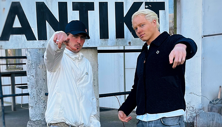 Två unga män poserar framför en vägg med texten Antikt.